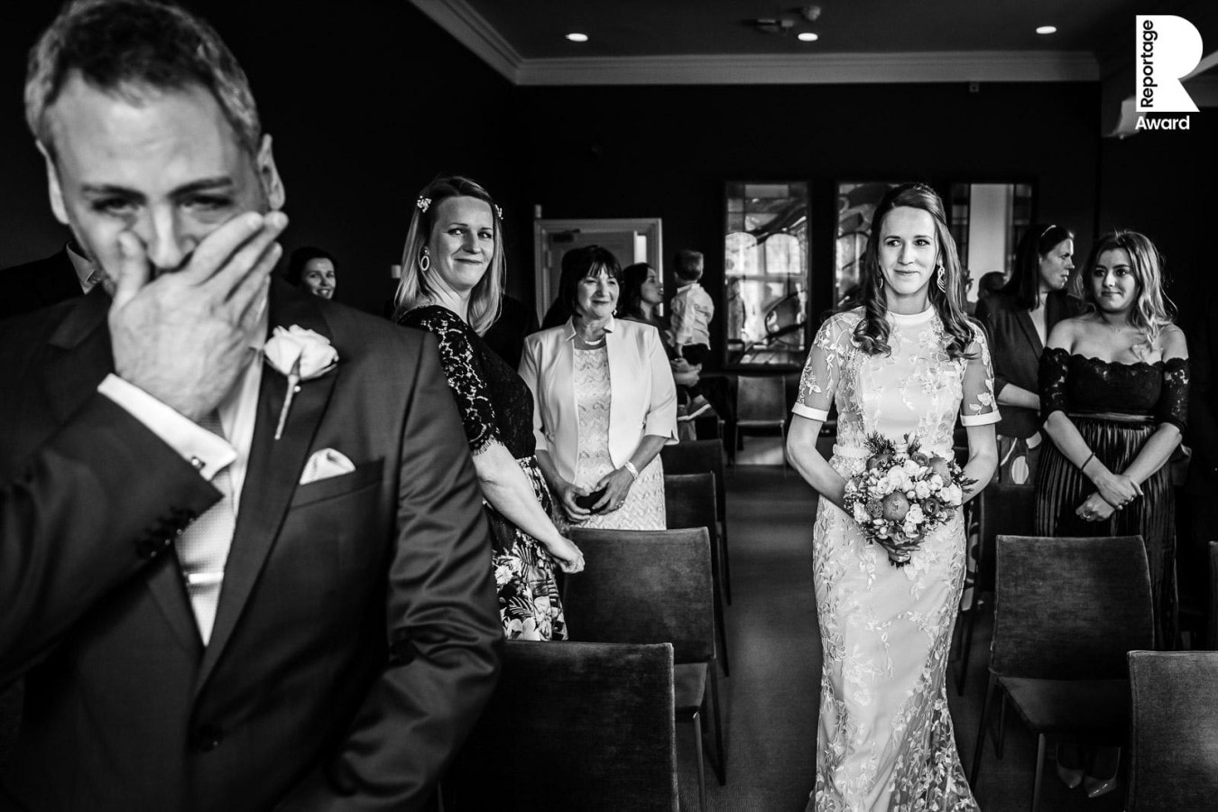 Bride walking down aisle, groom with face over mouth holding back emotions. B&W photo. This is Reportage Award June 2020