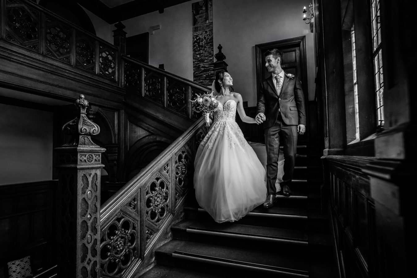 Black and white photo of wedding couple walking down the main staircase at Frensham Hall Wedding. Bride has a lovely flowing dress with a tight bodice and lace detail.